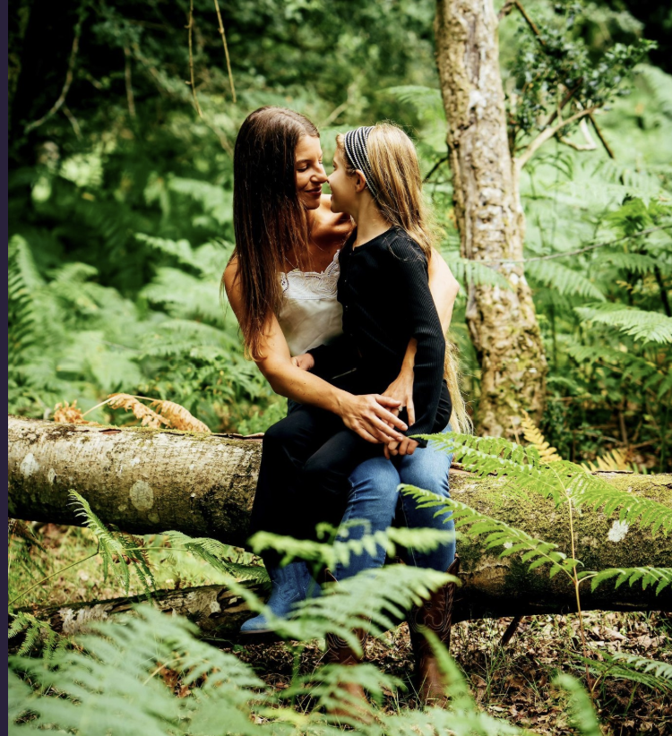 Two women sitting together on a mossy log in a sunlit forest, holding hands and smiling